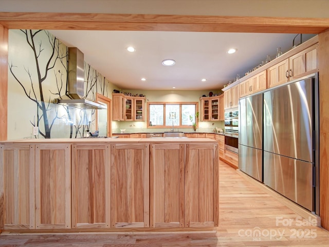 kitchen with glass insert cabinets, wall chimney exhaust hood, stainless steel fridge, and light brown cabinets