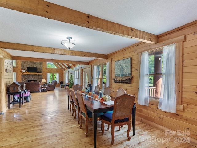 dining area featuring wood walls, a stone fireplace, a textured ceiling, beamed ceiling, and light hardwood / wood-style floors