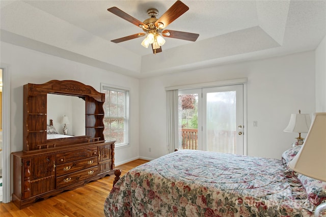 bedroom featuring access to outside, ceiling fan, a tray ceiling, and light wood-type flooring