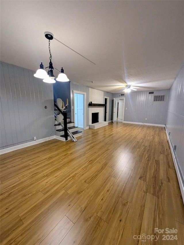 interior space featuring a large fireplace, wooden walls, ceiling fan with notable chandelier, and light wood-type flooring