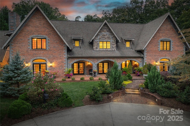 view of front facade featuring french doors, brick siding, a yard, roof with shingles, and stone siding