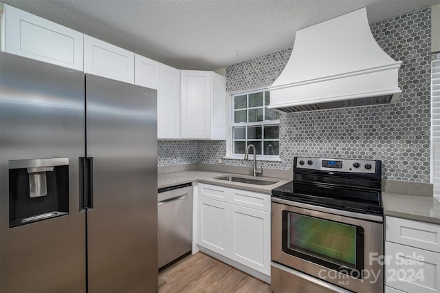 kitchen featuring appliances with stainless steel finishes, custom range hood, white cabinets, light wood-type flooring, and sink