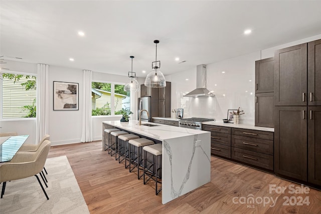 kitchen featuring light wood-type flooring, pendant lighting, an island with sink, a kitchen breakfast bar, and wall chimney exhaust hood