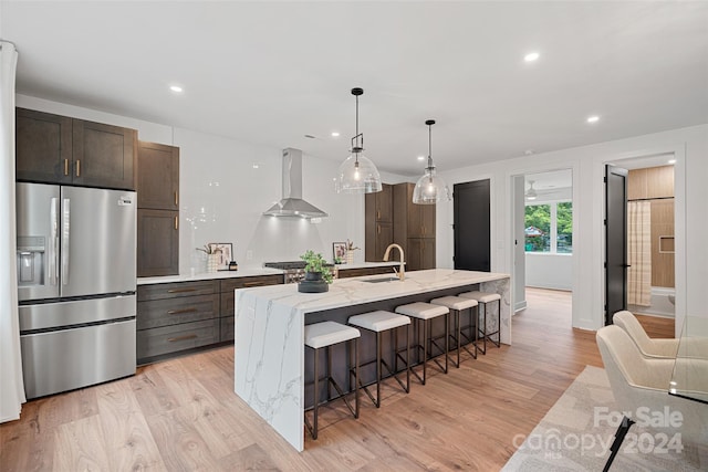 kitchen featuring a kitchen island with sink, stainless steel refrigerator with ice dispenser, wall chimney range hood, and light wood-type flooring