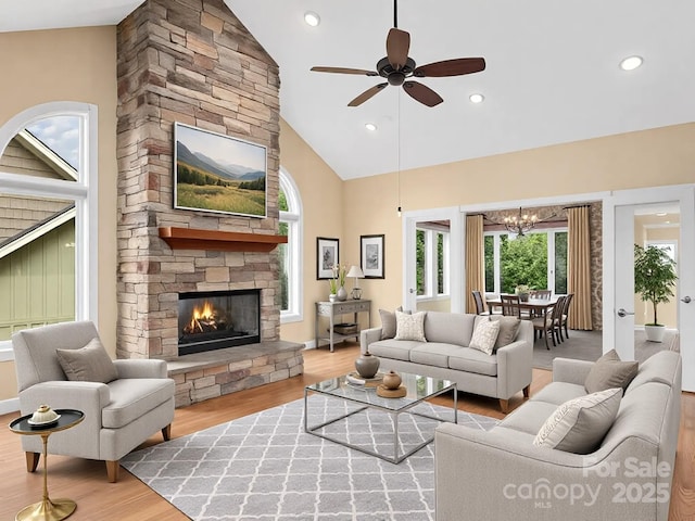 living room with ceiling fan with notable chandelier, a stone fireplace, high vaulted ceiling, and light wood-type flooring