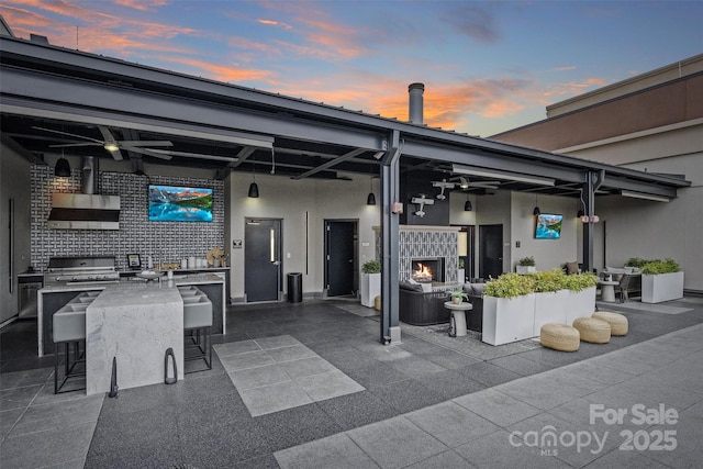 patio terrace at dusk with exterior kitchen, a bar, grilling area, and ceiling fan