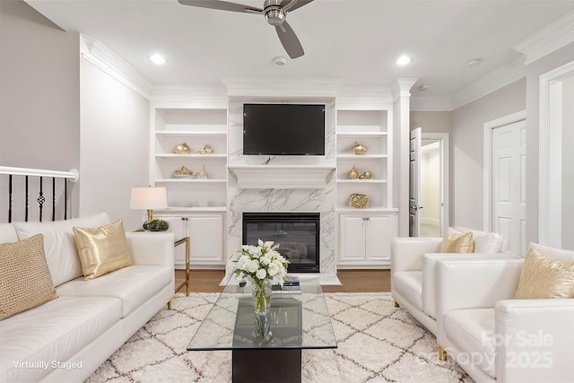 living room featuring built in shelves, wood-type flooring, ornamental molding, ceiling fan, and a high end fireplace