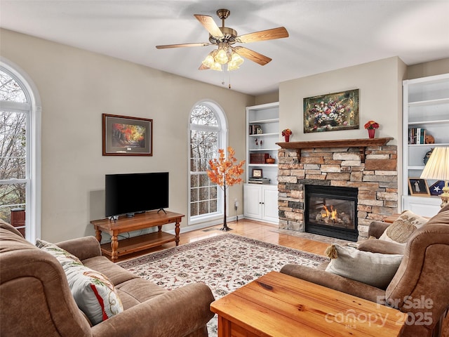 living room with built in shelves, a stone fireplace, plenty of natural light, and ceiling fan