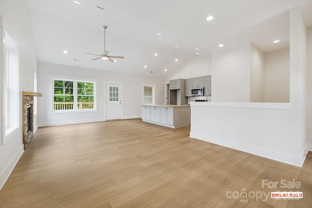 unfurnished living room featuring sink, light hardwood / wood-style flooring, ceiling fan, high vaulted ceiling, and a stone fireplace