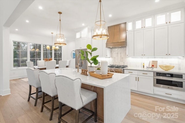 kitchen with stainless steel oven, an island with sink, custom exhaust hood, and white cabinets