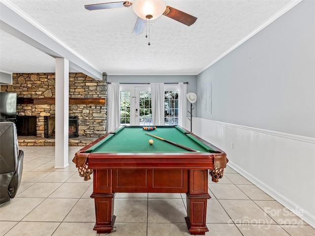 recreation room featuring ceiling fan, a stone fireplace, a textured ceiling, and ornamental molding