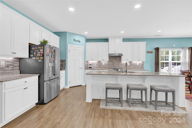 kitchen with light wood-type flooring, stainless steel fridge, a kitchen island with sink, white cabinetry, and light stone countertops