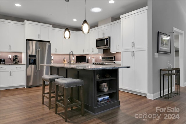 kitchen with white cabinets, dark stone counters, appliances with stainless steel finishes, decorative light fixtures, and a center island with sink