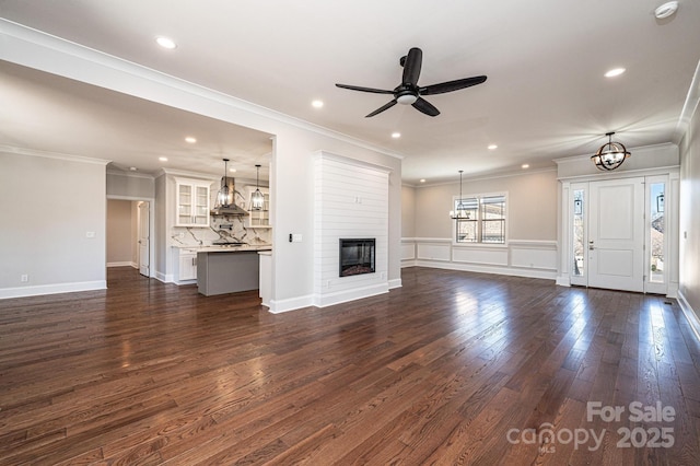 unfurnished living room with crown molding, ceiling fan with notable chandelier, dark wood-type flooring, and a glass covered fireplace