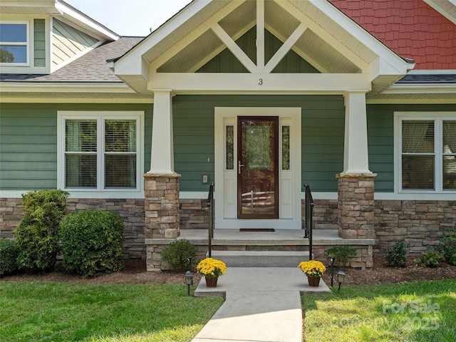 doorway to property featuring stone siding, covered porch, and roof with shingles