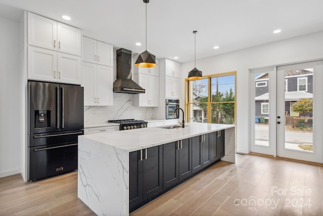 kitchen featuring wall chimney exhaust hood, hanging light fixtures, stainless steel appliances, a center island with sink, and white cabinets