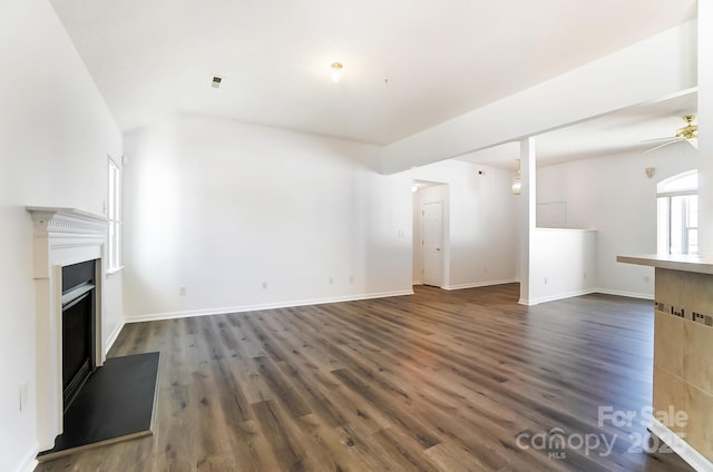 unfurnished living room with a ceiling fan, baseboards, dark wood-type flooring, and a fireplace with raised hearth