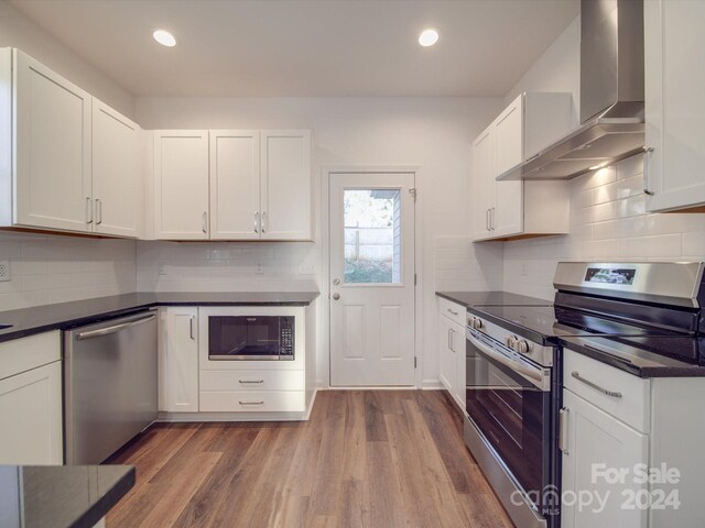 kitchen with dark countertops, wall chimney range hood, appliances with stainless steel finishes, and white cabinetry