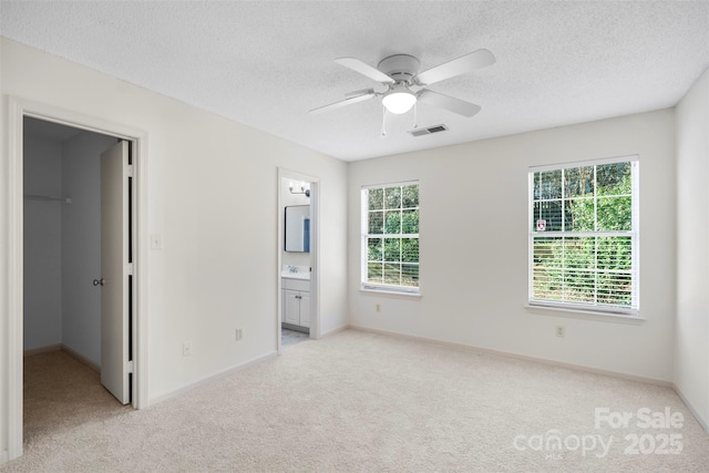 unfurnished bedroom with ensuite bath, a spacious closet, ceiling fan, light carpet, and a textured ceiling