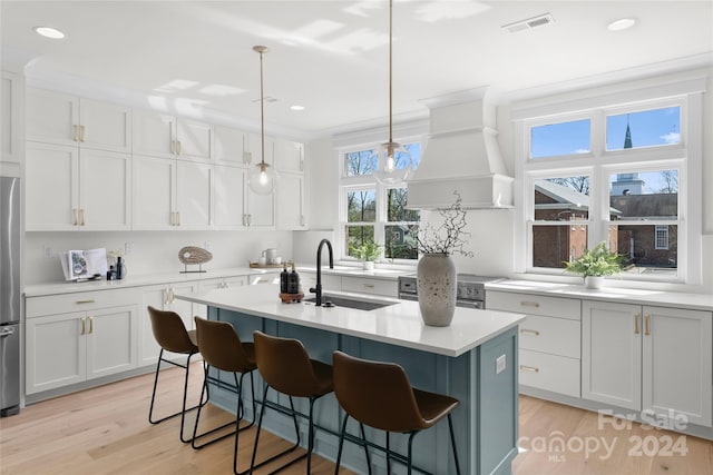 kitchen with pendant lighting, premium range hood, a kitchen island with sink, white cabinets, and light hardwood / wood-style flooring