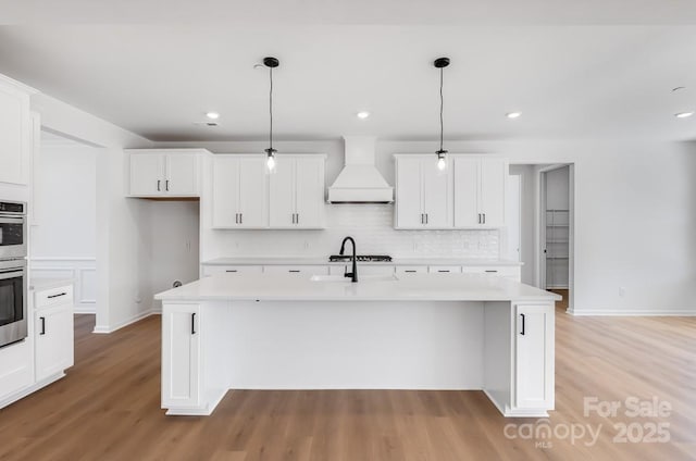 kitchen with light countertops, custom range hood, decorative light fixtures, and a center island with sink