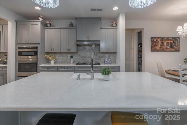 kitchen with stainless steel double oven, tasteful backsplash, a notable chandelier, gray cabinetry, and a center island with sink