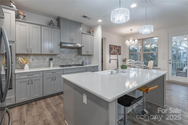 kitchen with pendant lighting, appliances with stainless steel finishes, backsplash, a kitchen island with sink, and a notable chandelier