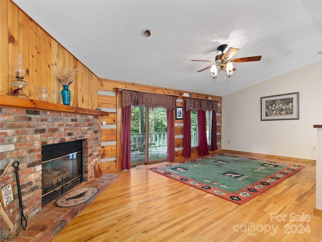 living room featuring ceiling fan, wood walls, a brick fireplace, a textured ceiling, and light hardwood / wood-style floors