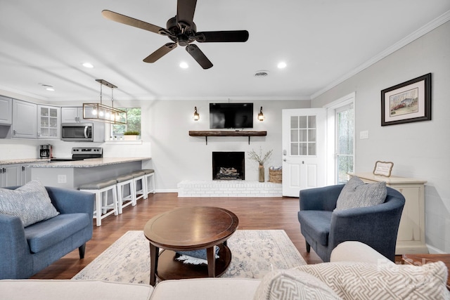 living room with a brick fireplace, ceiling fan, dark wood-type flooring, and crown molding