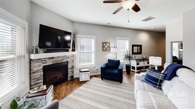living room with ceiling fan, a stone fireplace, visible vents, wood finished floors, and baseboards