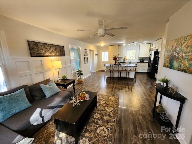 living room featuring a decorative wall, dark wood-type flooring, a ceiling fan, wainscoting, and rail lighting