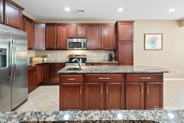 kitchen featuring backsplash, appliances with stainless steel finishes, light tile patterned floors, light stone countertops, and a kitchen island with sink