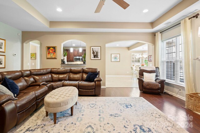 living room featuring ceiling fan with notable chandelier, wood-type flooring, and a tray ceiling