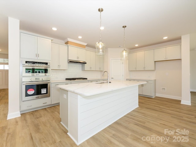 kitchen featuring light wood-style floors, an island with sink, double oven, gas stovetop, and light stone counters