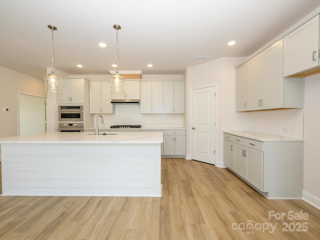 kitchen with light wood-style flooring, stainless steel double oven, a kitchen island with sink, a sink, and decorative backsplash