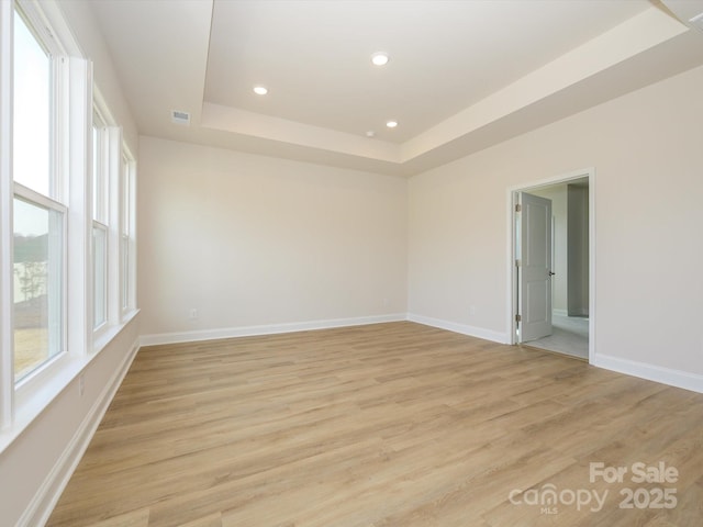 empty room with light wood-style floors, visible vents, baseboards, and a raised ceiling
