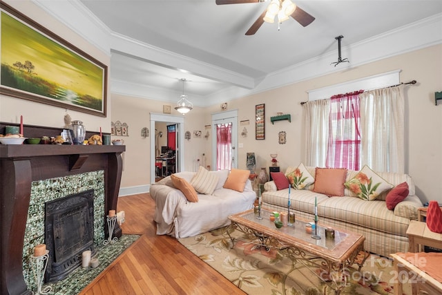 living room with ceiling fan, beamed ceiling, wood-type flooring, a tiled fireplace, and crown molding