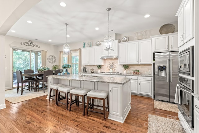 kitchen with white cabinets, hanging light fixtures, wood-type flooring, a kitchen island with sink, and appliances with stainless steel finishes