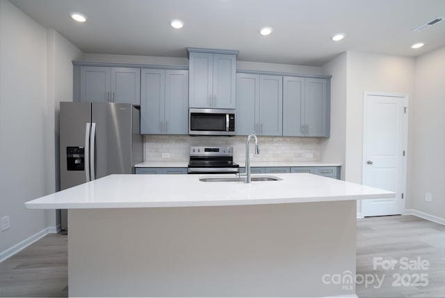 kitchen featuring sink, light wood-type flooring, appliances with stainless steel finishes, an island with sink, and decorative backsplash