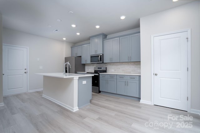 kitchen featuring gray cabinets, appliances with stainless steel finishes, an island with sink, backsplash, and light hardwood / wood-style floors