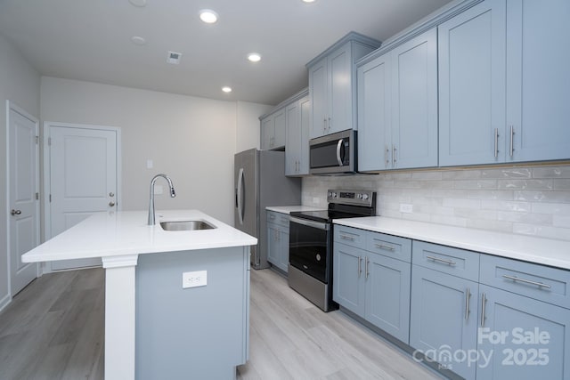 kitchen with appliances with stainless steel finishes, tasteful backsplash, an island with sink, sink, and light hardwood / wood-style floors