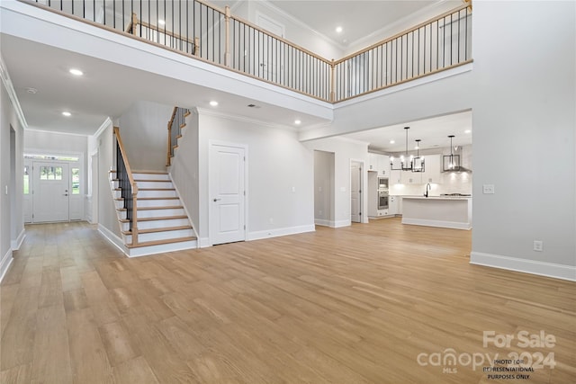 unfurnished living room featuring an inviting chandelier, light wood-style flooring, a towering ceiling, stairs, and baseboards