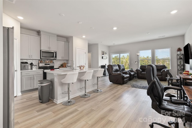 kitchen featuring a center island with sink, backsplash, stainless steel appliances, light wood-type flooring, and a kitchen bar