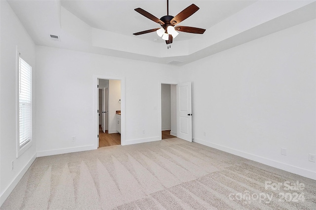 unfurnished bedroom featuring a raised ceiling, ceiling fan, light colored carpet, and ensuite bath