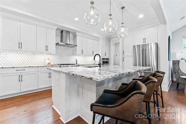 kitchen with wall chimney range hood, an island with sink, sink, white cabinets, and appliances with stainless steel finishes