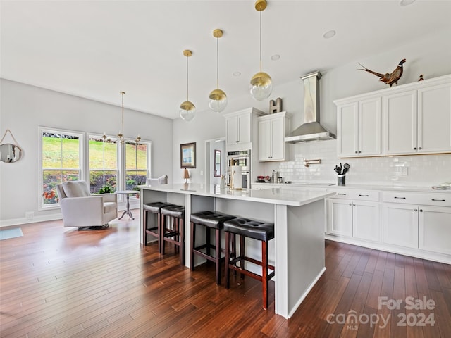 kitchen with dark hardwood / wood-style flooring, a center island with sink, pendant lighting, wall chimney range hood, and white cabinetry