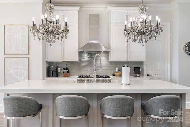 kitchen with a kitchen bar, a kitchen island with sink, wall chimney range hood, backsplash, and white cabinetry