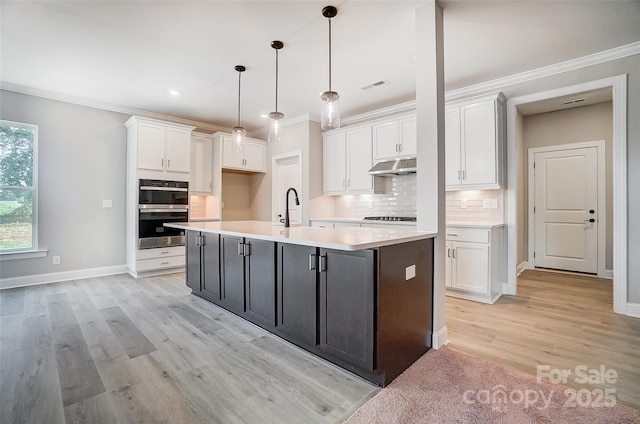 kitchen with white cabinetry, hanging light fixtures, stainless steel double oven, backsplash, and a kitchen island with sink