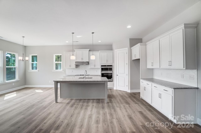 kitchen featuring white cabinets, a kitchen island with sink, light hardwood / wood-style flooring, stone counters, and double oven