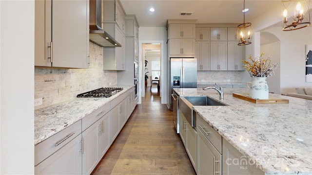 kitchen with light stone countertops, sink, wall chimney range hood, pendant lighting, and appliances with stainless steel finishes